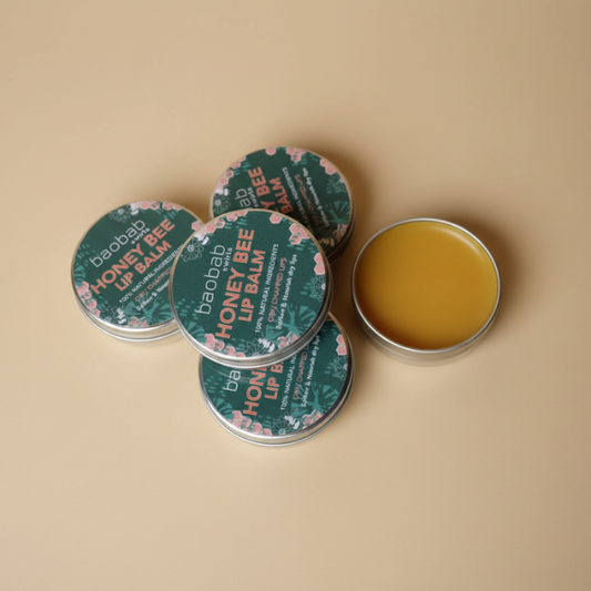 Four round metal tins of baobab swirls Honey Bee Lip Balm arranged on a warm beige background, with one tin open to show the golden-yellow balm inside.